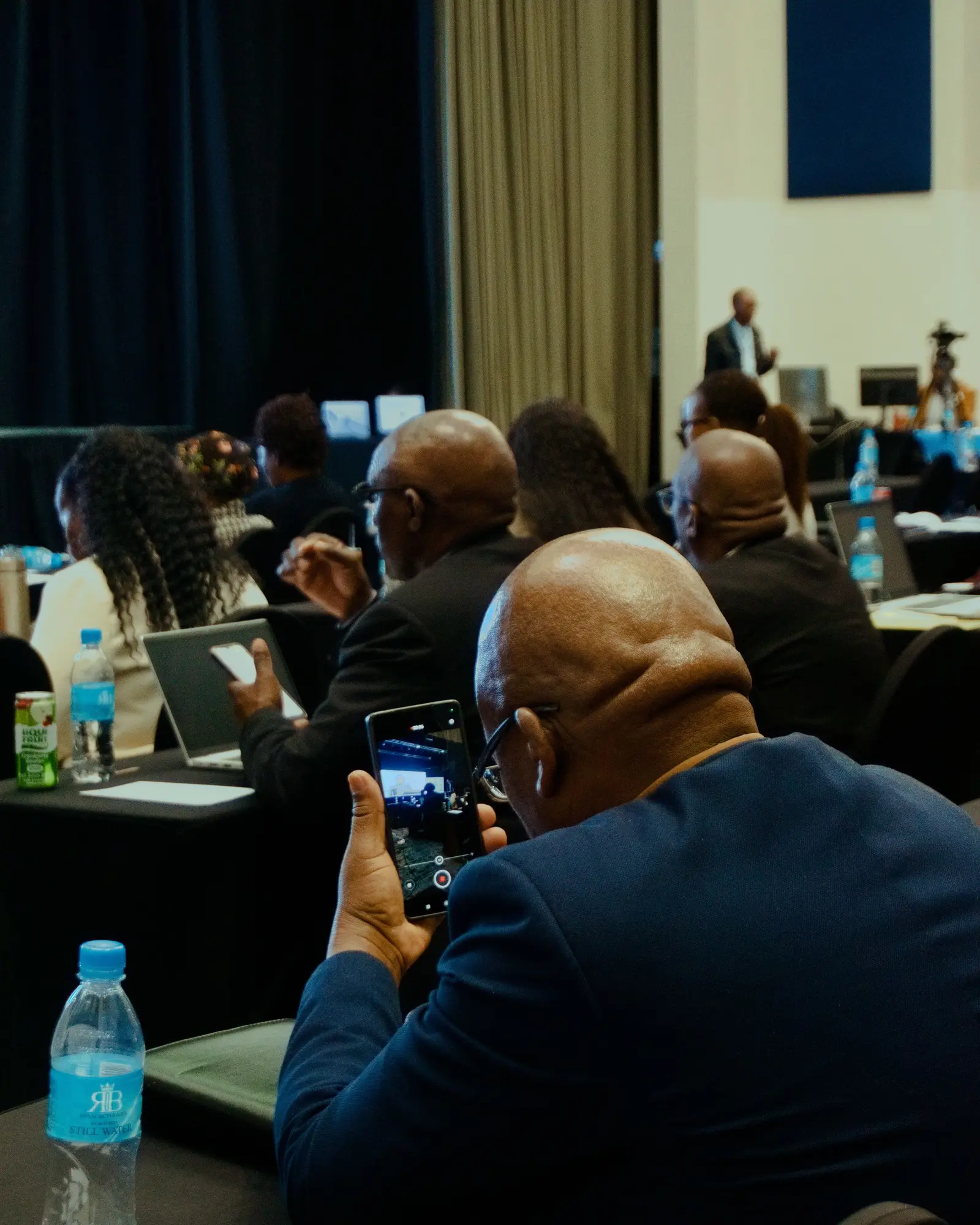 Seated audience at a business conference following a speaker on stage.